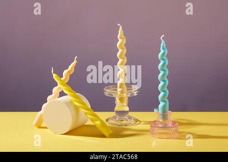 Two twisted candles are placed on glass candlesticks, two others are leaned next to the white podium. Yellow and purple background. Birthday with cand Stock Photo