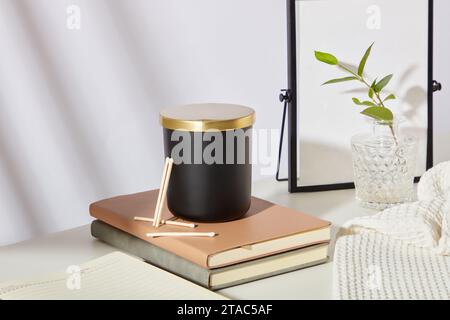 A jar of unlabeled scented candles is placed on top of two notebooks. A mirror, glass flower vase and cloth towels are displayed on the table. Delicat Stock Photo