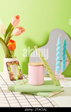 Tulips are placed in a porcelain vase. A jar of scented candles is displayed on the table with two notebooks. Colored candles and a mirror are placed Stock Photo