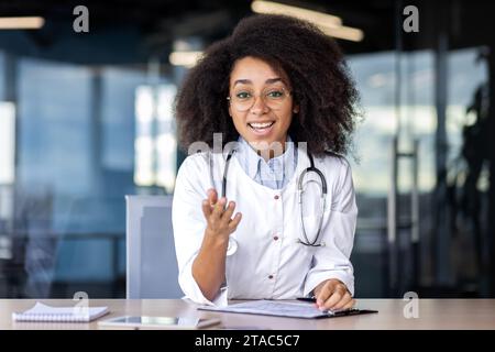 Web camera view, screen, female doctor talking remotely with patients, consulting, happy looking at camera, gesturing with hands, female doctor in white medical coat inside clinic office. Stock Photo