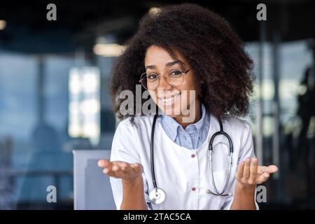 Web camera view, screen, female doctor talking remotely with patients, consulting, happy looking at camera, gesturing with hands, female doctor in white medical coat inside clinic office. Stock Photo