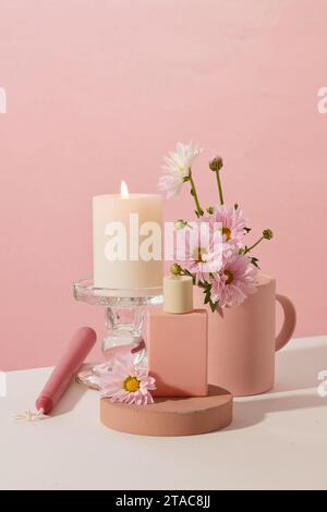 A burning candle is placed on a glass candlestick, an unlabeled perfume bottle is placed on a wooden podium and fresh flowers are on a pink and white Stock Photo