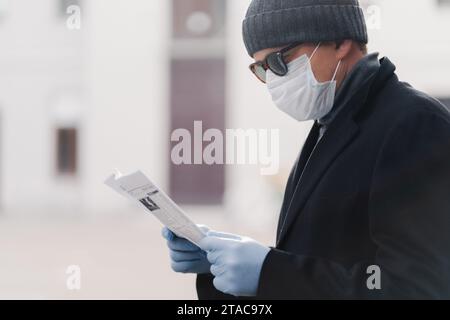Man in winter attire reading a newspaper outdoors, wearing a mask and gloves for safety Stock Photo