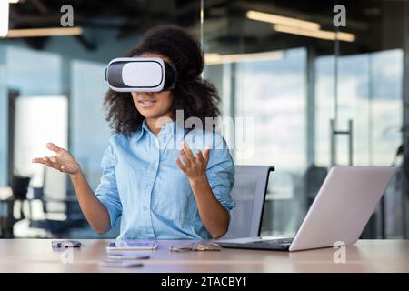 Businesswoman at workplace wearing virtual reality glasses communicating remotely in video simulator, female office worker working with laptop, remote meeting. Stock Photo