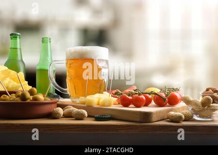 Freshly prepared appetizer on wooden bench with beer mug, cheese, peanuts, olives, cherry tomatoes, and other snacks on cutting board in the backgroun Stock Photo