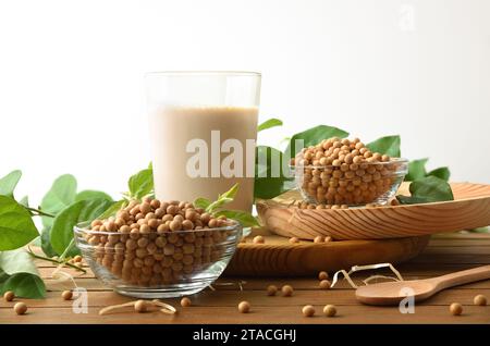 Background with bowls full of dry soy beans and glass with soy drink on wooden table with leaves and isolated white background. Front view. Stock Photo