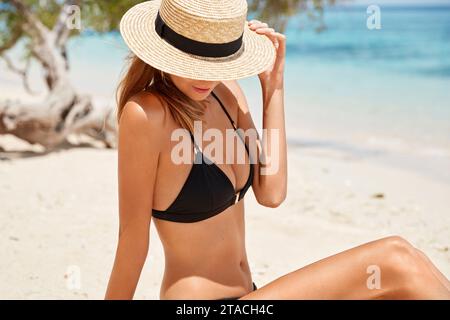 Outdoor shot of slim female model in black bikini and summer hat, sits on sandy beach alone, poses against beautiful ocean view, enjoys summer time. A Stock Photo