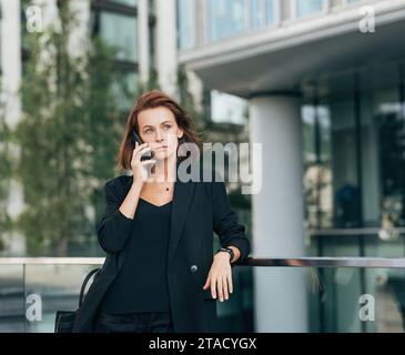 Confident middle-aged businesswoman with ginger hair leaning on railing talking on mobile phone outdoors Stock Photo