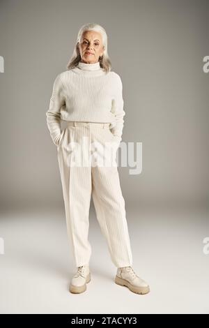attractive and grey haired middle aged woman in white winter outfit standing with hands in pockets Stock Photo