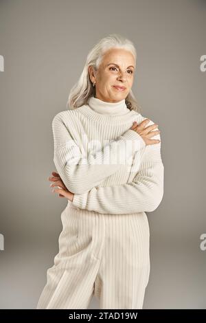 charming and grey haired middle aged woman in white winter sweater standing on grey background Stock Photo