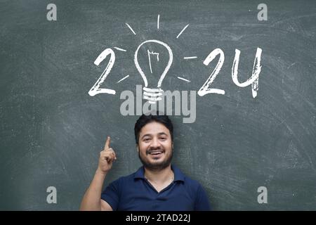 Indian/Asian young man pointing up while standing against blackboard with chalk drawing of light bulb and new year 2024 Stock Photo