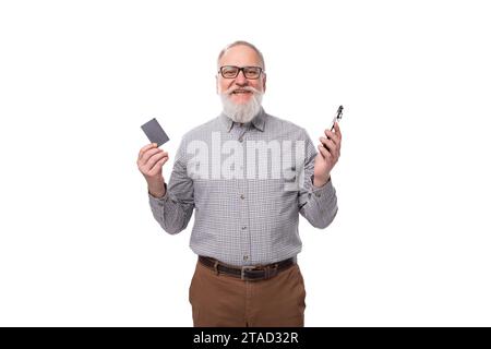 modern smart gray-haired mature businessman with beard and mustache in shirt and trousers holds credit card mockup and smartphone Stock Photo