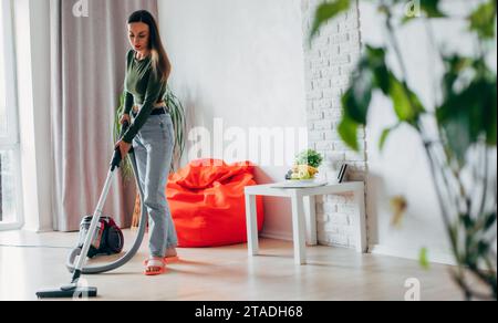 Full-length photo of beautiful young woman is using a vacuum cleaner while cleaning floor at home on living room Stock Photo