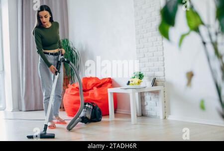Full-length photo of beautiful young woman is using a vacuum cleaner while cleaning floor at home on living room Stock Photo