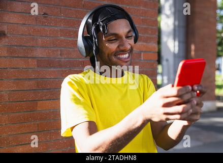 Young urban black man with a durag, smiling cheerfully, wearing headphones, and interacting with his mobile phone Stock Photo
