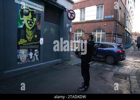 A man photographs an image of Shane MacGowan, lead singer of The Pogues, on a building in the Temple area of Dublin. Shane MacGowan has died aged 65, his wife has announced in a post on Instagram. Picture date: Thursday November 30, 2023. Stock Photo