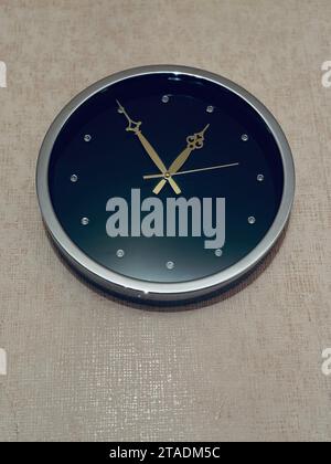 A black and gold wall clock hangs on a tan wall background Stock Photo