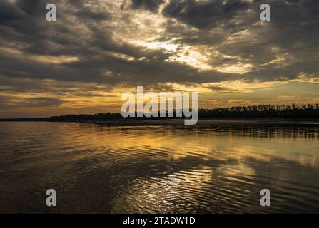 Clouds hide setting sun giving golden glow across the calm Mississippi River in Missouri Stock Photo