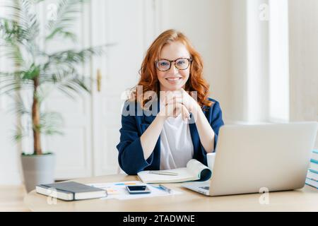Smiling redhead woman with glasses working on a laptop at a bright home office Stock Photo