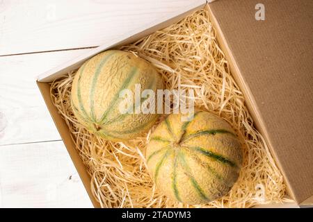 Two fragrant organic melons in a paper box with shavings, close-up, on a white wooden table, top view. Stock Photo