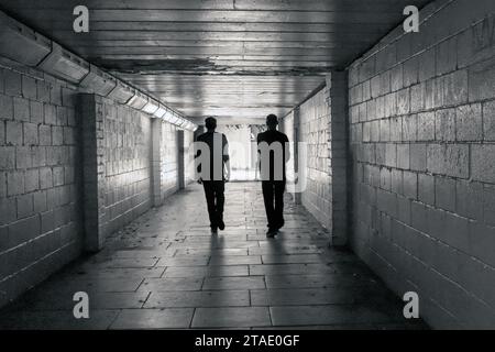 Monochrome image of silhouette of two men walking in a dark pedestrian underpass towards exit and daylight. Stock Photo