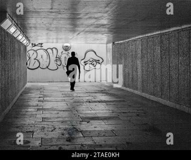 Black and white image of silhouette of a person walking in a dark pedestrian underpass with graffiti on wall behind him. Stock Photo