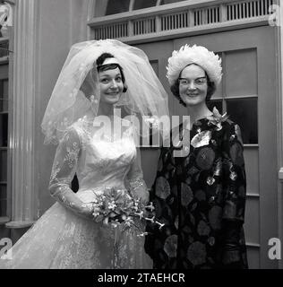 1960s, historical, wedding, inside a room, a young bride in her wedding dress and bridal hair net, standing with her proud mother for a photo, who is wearing spectacles of the era and a fancy hat, England, UK. Stock Photo