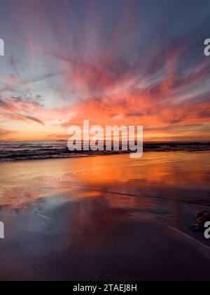 Low angle view of a dramatic beach sunset with vibrant clouds reflecting in the wet sand on shore as waves wash up in the horizon. Stock Photo