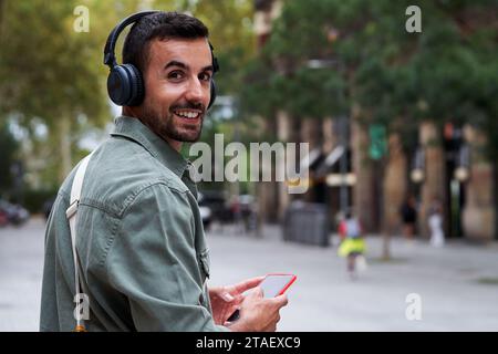 Portrait of a young man walking outside in the street, using phone to listen music, turns to camera. Stock Photo