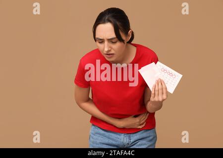 Young woman holding pad and calendar with marked days of menstruation on brown background Stock Photo