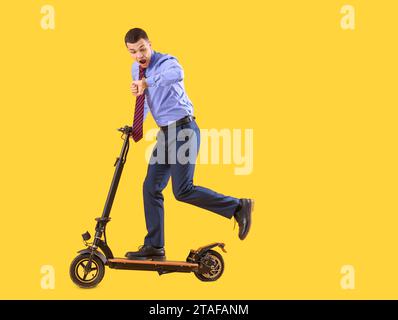 Shocked young businessman with electric scooter looking at wristwatch on yellow background Stock Photo