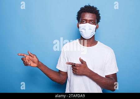 Afro haired black man in portrait facing the camera and gesturing with his hands to the right. African American guy with a face mask marketing a product while motioning with his arms. Stock Photo