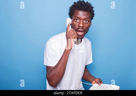 Surprised young black man with afro hair speaks on a telephone in a studio. African american guy holds landline phone in hand against blue background, expressing shock during conversation. Stock Photo