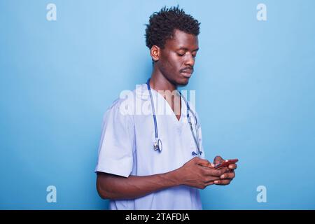 Portrait of African American medical professional stands against blue background holding modern cell phone. Wearing blue scrubs is male nurse using smartphone to provide healthcare. Stock Photo