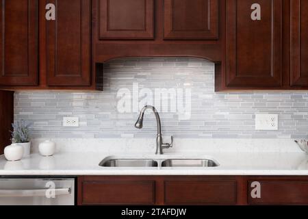A kitchen faucet detail with a glass tiled backsplash, dark wood cabinets, and a white marble countertop. Stock Photo