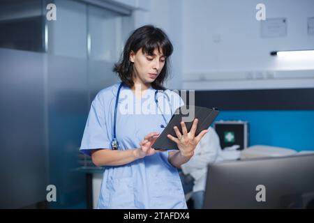 Detailed image of a caucasian woman in blue scrubs standing in a clinic office using a tablet for medical research. Female nurse utilizing a digital device to review patient appointments. Stock Photo