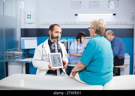 During medical checkup, healthcare specialist shows picture of human skeleton on tablet to elderly patient. Male doctor holds up gadget that displays examination of bones and diagnoses osteopathy. Stock Photo