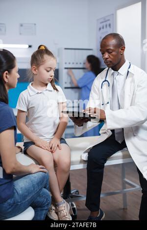 In clinic room, a doctor explains treatment to a young caucasian girl while a nurse works in background. The focus is on healthcare and medicine, with a stethoscope and digital device visible. Stock Photo