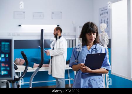Physiotherapy office with portrait of nurse practitioner standing, checking her paperwork. Medical physician getting ready to assist patients with physical rehabilitation exercises. Stock Photo