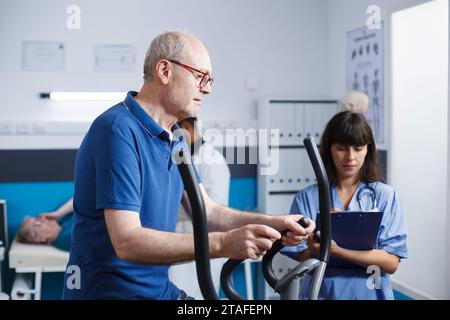 Elderly man is accompanied by female physician wearing blue scrubs as he receives physical therapy on stationary bike. Old patient is receiving rehabilitative care from medical practitioner. Stock Photo
