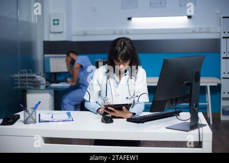 Front-view of female physician using a tablet in clinic office to analyze healthcare data. Caucasian doctor in lab coat, concentrated on reviewing patient medical information on her digital device. Stock Photo