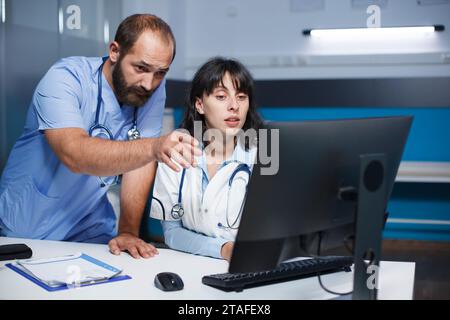 Male nurse in blue scrubs pointing toward a pc monitor while talking to a doctor wearing a lab coat. Medical workers are seen using a computer and having a deep discussion on healthcare. Stock Photo