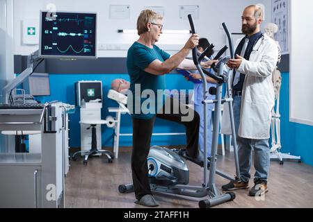 Elderly woman uses electric bicycle for physical rehabilitation after being hurt. Senior female patient using a stationary bike for gymnastics while the doctor assists with physiotherapy. Stock Photo