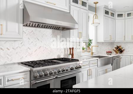 A kitchen detail with grey cabinets, a herringbone tile backsplash, stainless steel apron sink, and a gold hardware and faucet. No brands or logos. Stock Photo