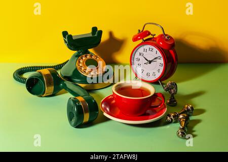 Red cup of tea, alarm clock, vintage telephone and chessmen on light green table against yellow background Stock Photo