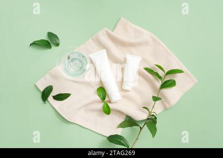 A glass of water and two unlabeled cosmetic tubes are displayed on a towel spread in the middle. Green tea leaves are decorated around. Green tea cont Stock Photo