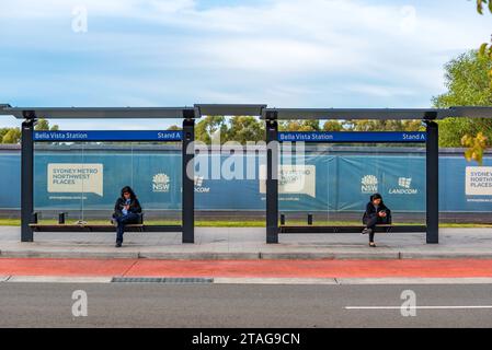 Two women people sitting separately looking at their phones at a bus stop near Bella Vista Metro Station in north west Sydney, Australia Stock Photo