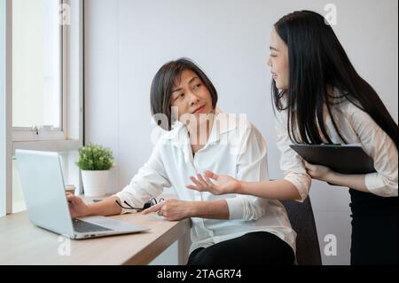 A beautiful young Asian businesswoman is talking with a senior female colleague in the office, sharing her ideas and opinions. Stock Photo