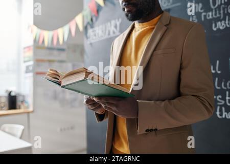 African American teacher standing near the blackboard and reading textbook during lesson Stock Photo