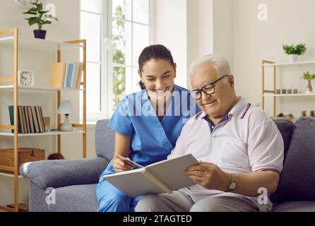 Friendly female nurse and older man are looking at photos from his family photo album together. Stock Photo